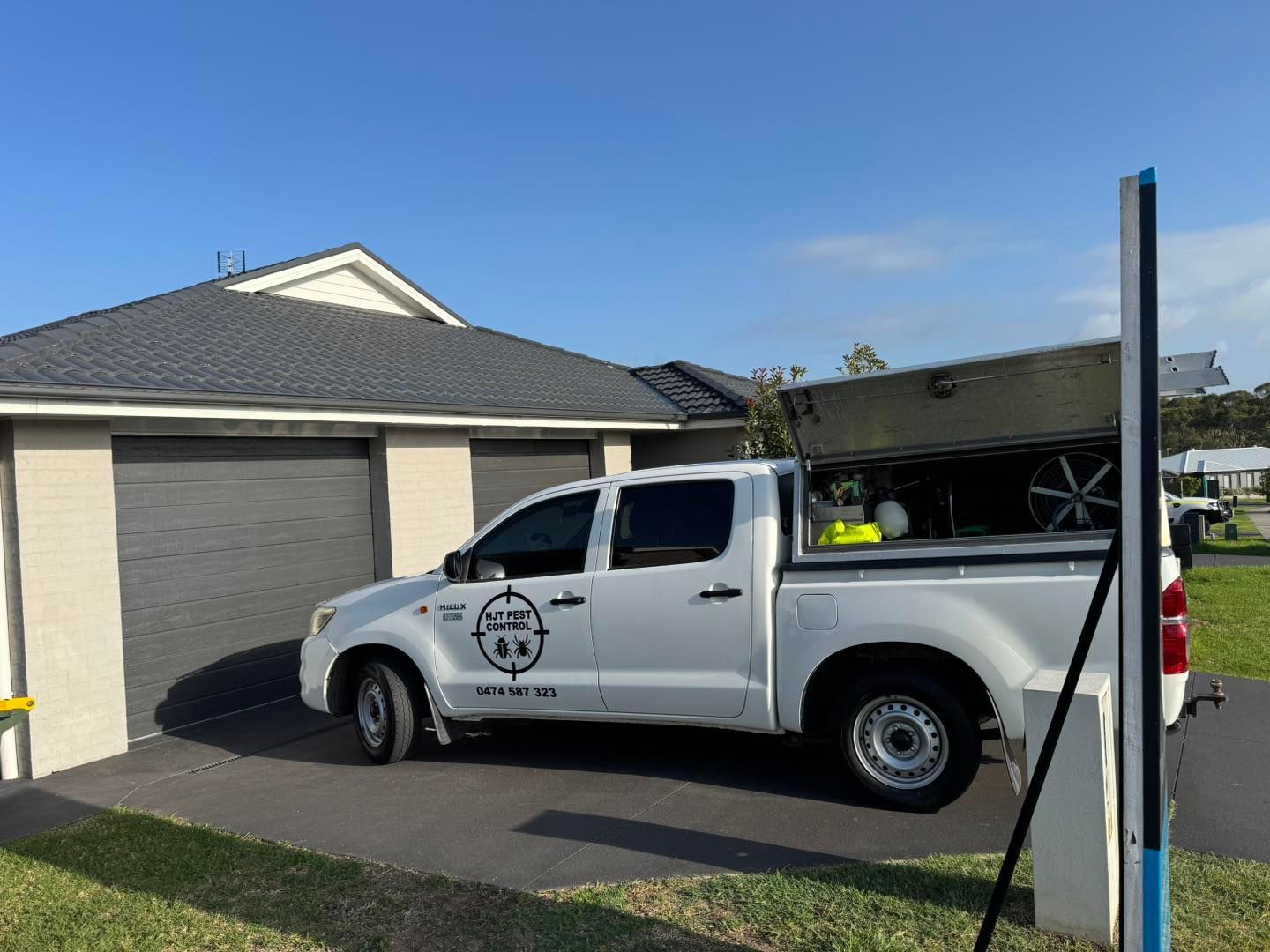 A white work truck with an open tool canopy parked in the driveway of a single-story brick house with a gray garage door.