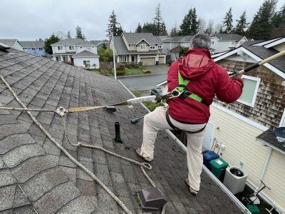 Worker in a red jacket cleaning a shingled roof with a safety harness, overlooking a suburban neighborhood.