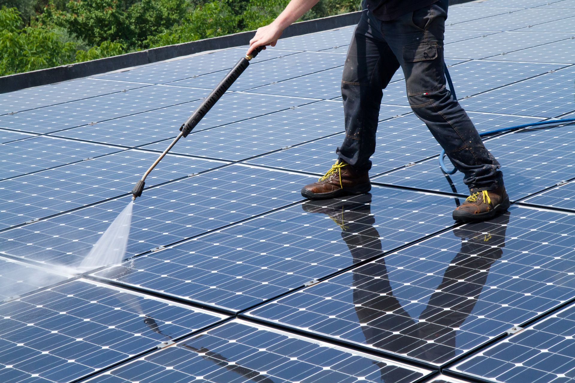 Worker pressure-washing solar panels on a rooftop in bright sunlight.