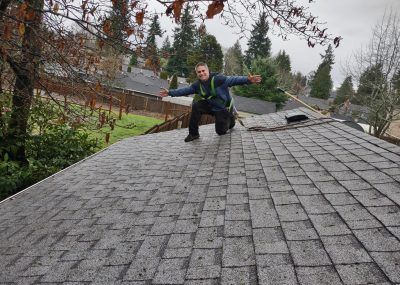 Person balancing on a steep gray shingled roof in a suburban neighborhood with trees and houses nearby.