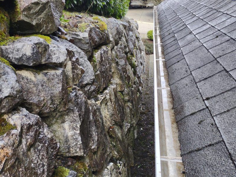 Stone retaining wall beside a gray shingle roof with a narrow drainage channel between them.