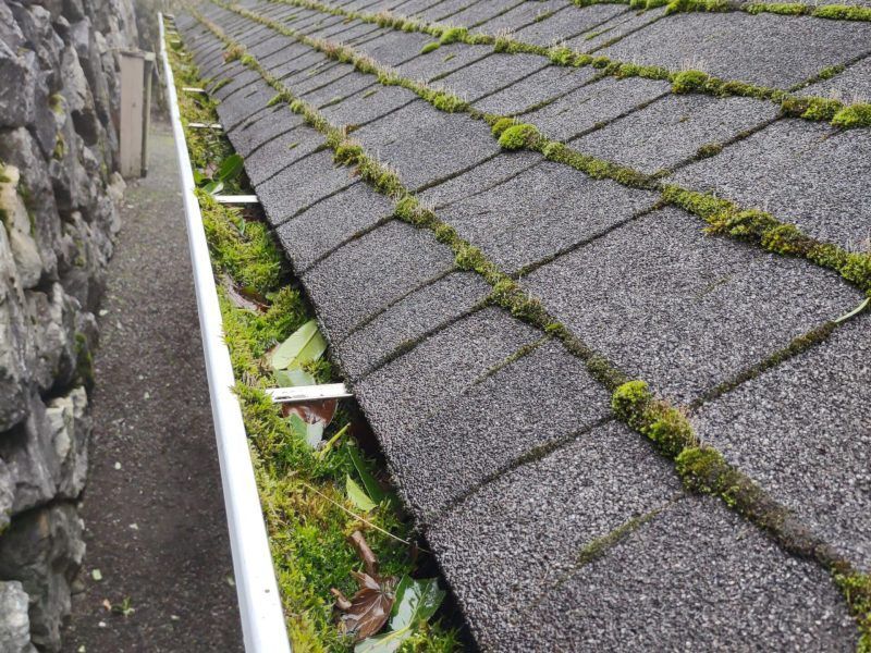 Moss-covered shingled roof beside a stone wall and white gutter along a narrow channel.