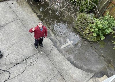 Person in a red jacket standing on a concrete path, looking up and waving near a garden wall.