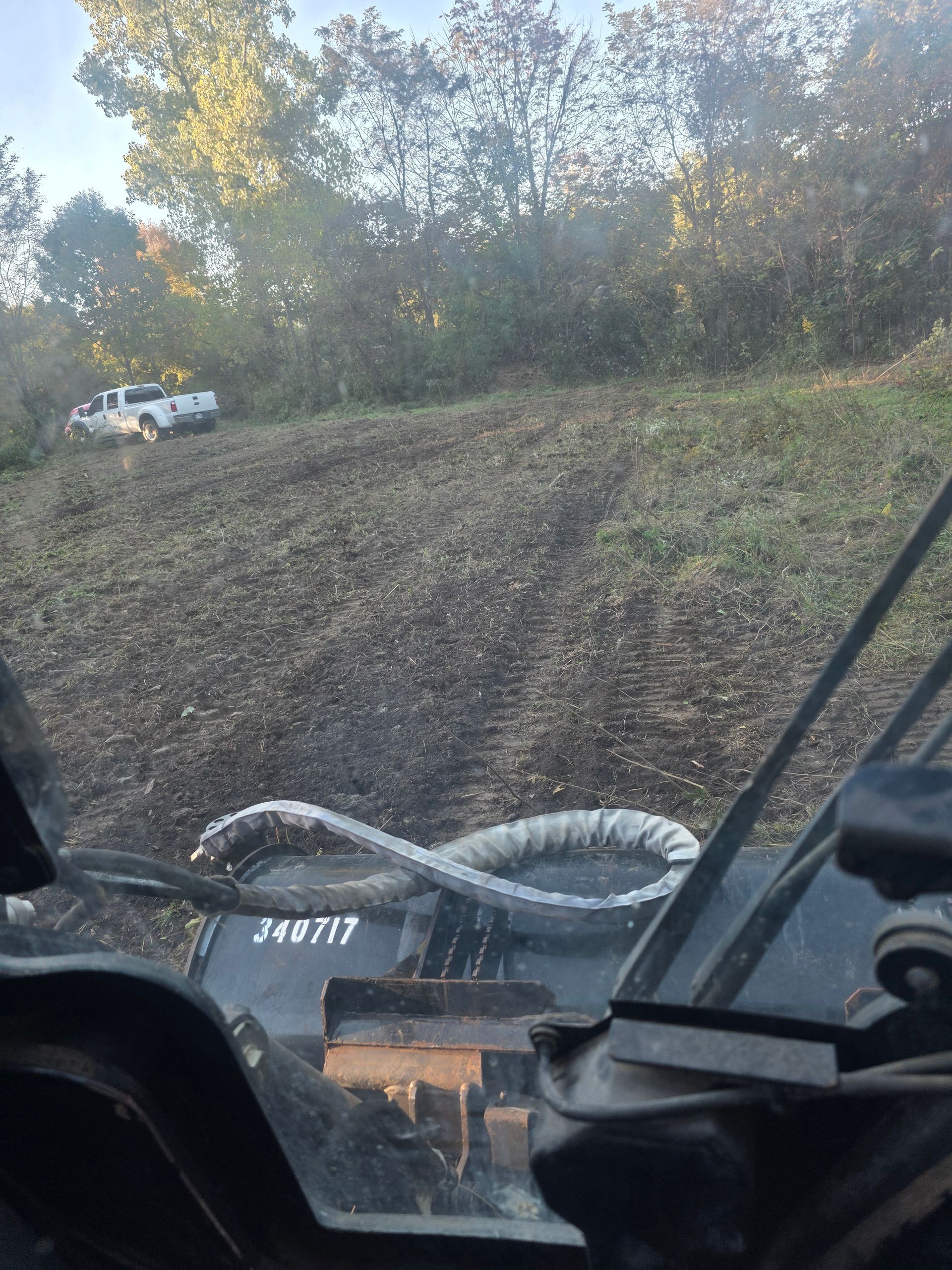 View from a vehicle cab over a muddy field with a white truck near the tree line.