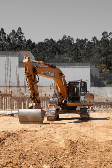 Orange excavator on a dirt construction site near shipping containers and trees.