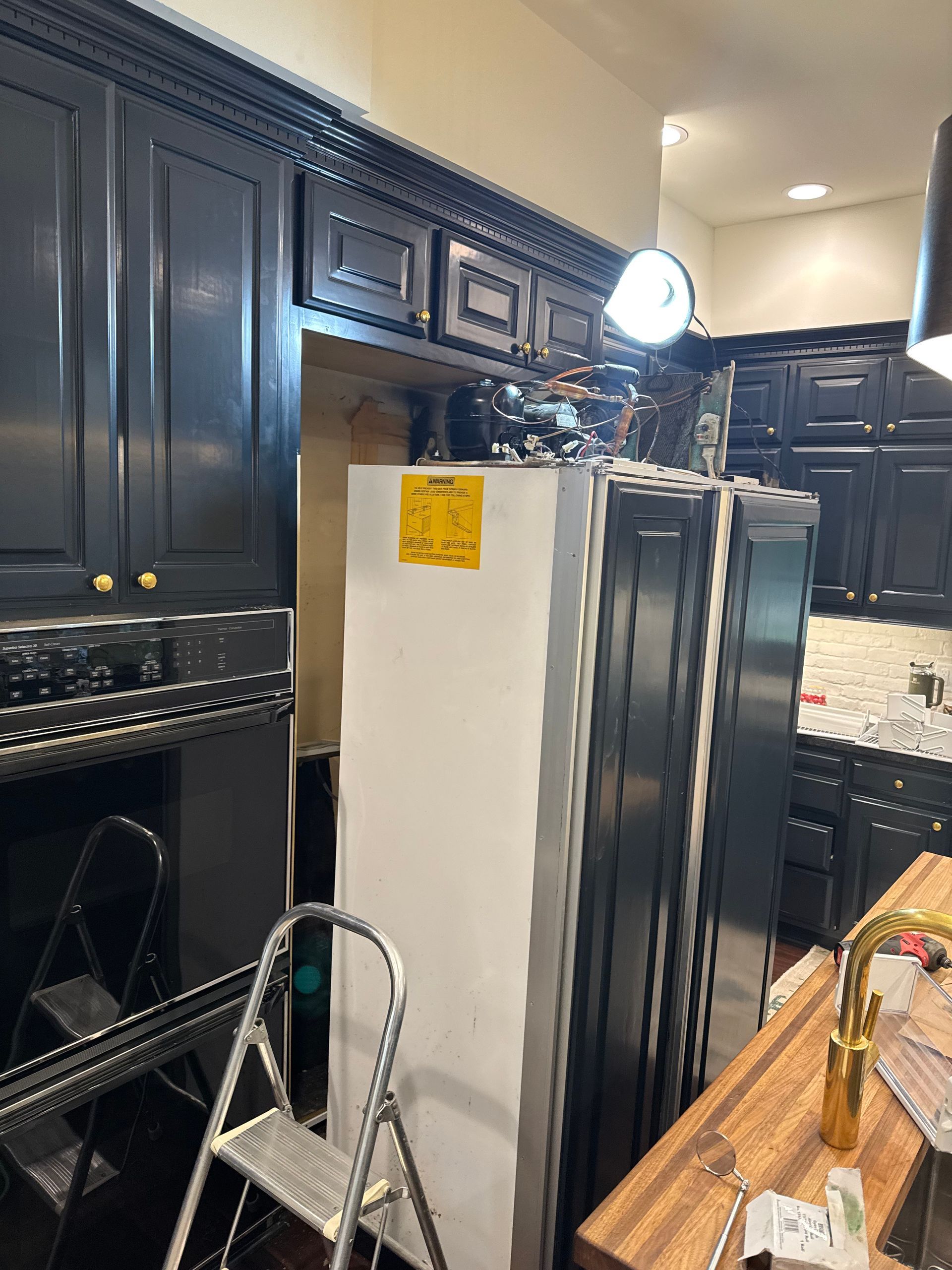 Refrigerator being installed in a kitchen. Black cabinets, oven, and a ladder are also present.