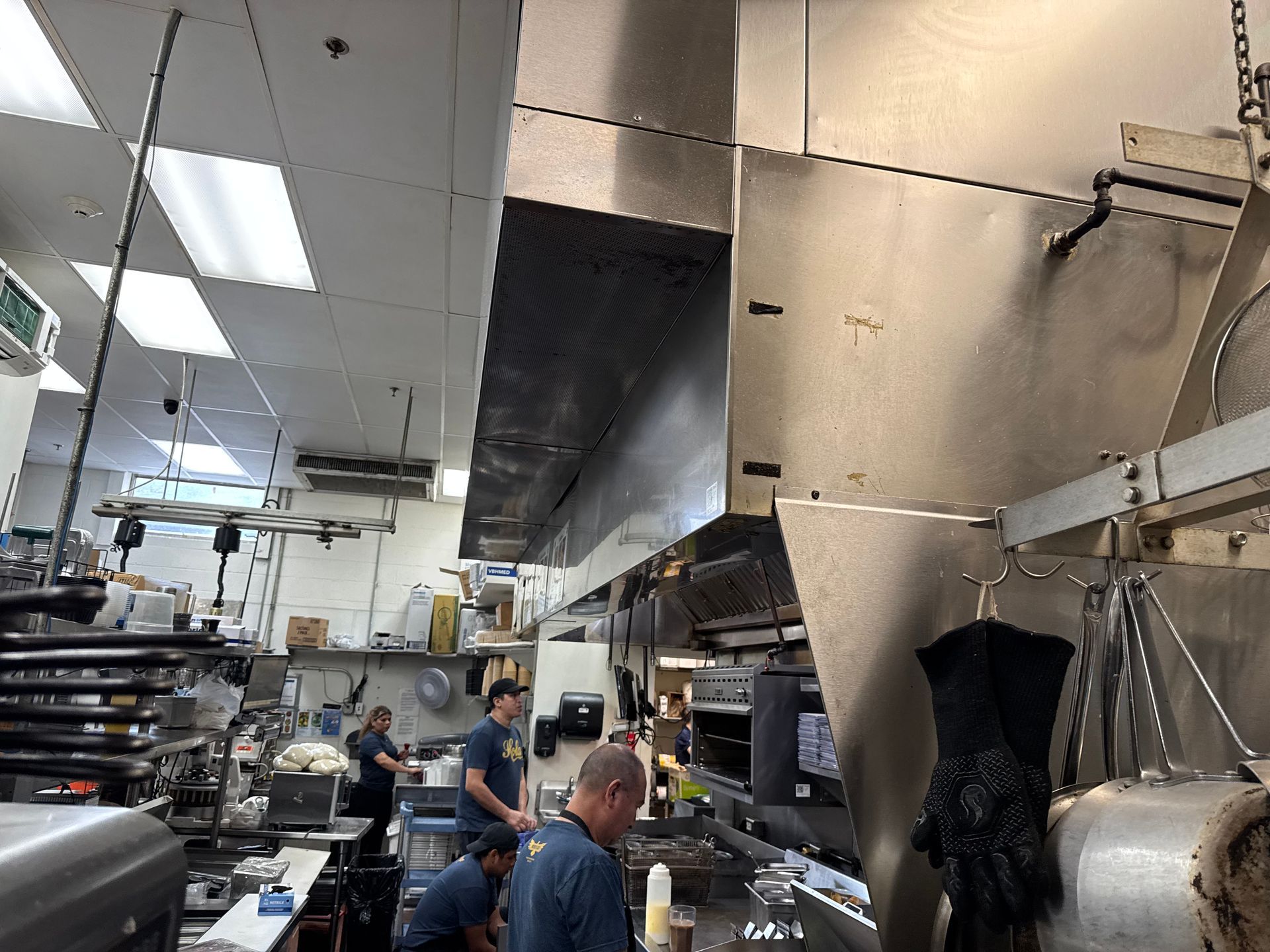 Commercial kitchen with cooks preparing food. Stainless steel hood, bright lights.