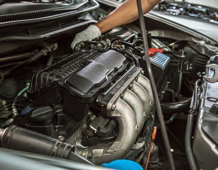 Open car engine bay with a hand inspecting the black engine cover and silver exhaust manifold.