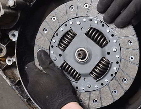 Gloved hands holding a clutch disc with springs, showing a worn automotive part on a workbench.