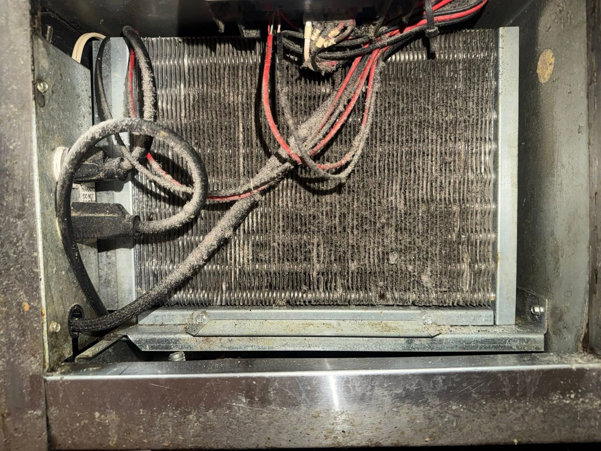 Dusty appliance compartment with a dirty coil, wires, and a metal fan housing inside.