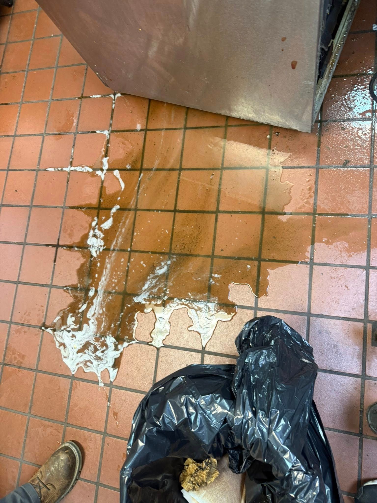 Spilled brown liquid on a tiled floor beside a black trash bag and a metal door