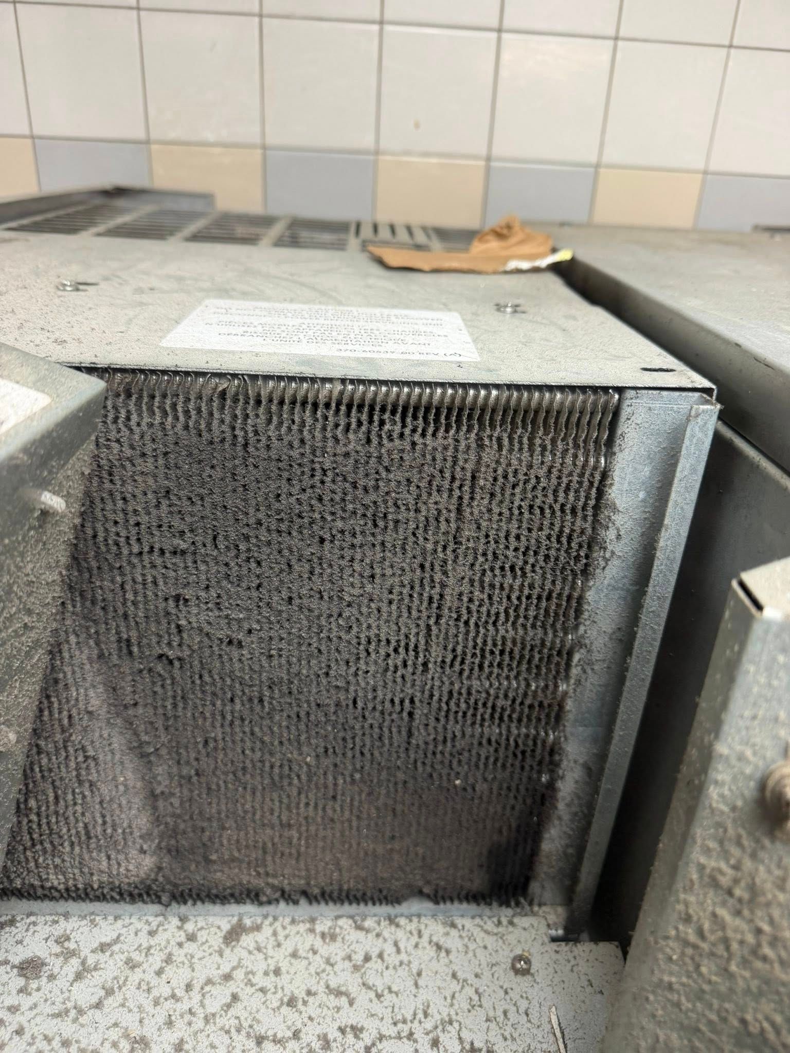 Dirty metal grease filter in a kitchen, leaning against a tiled wall.