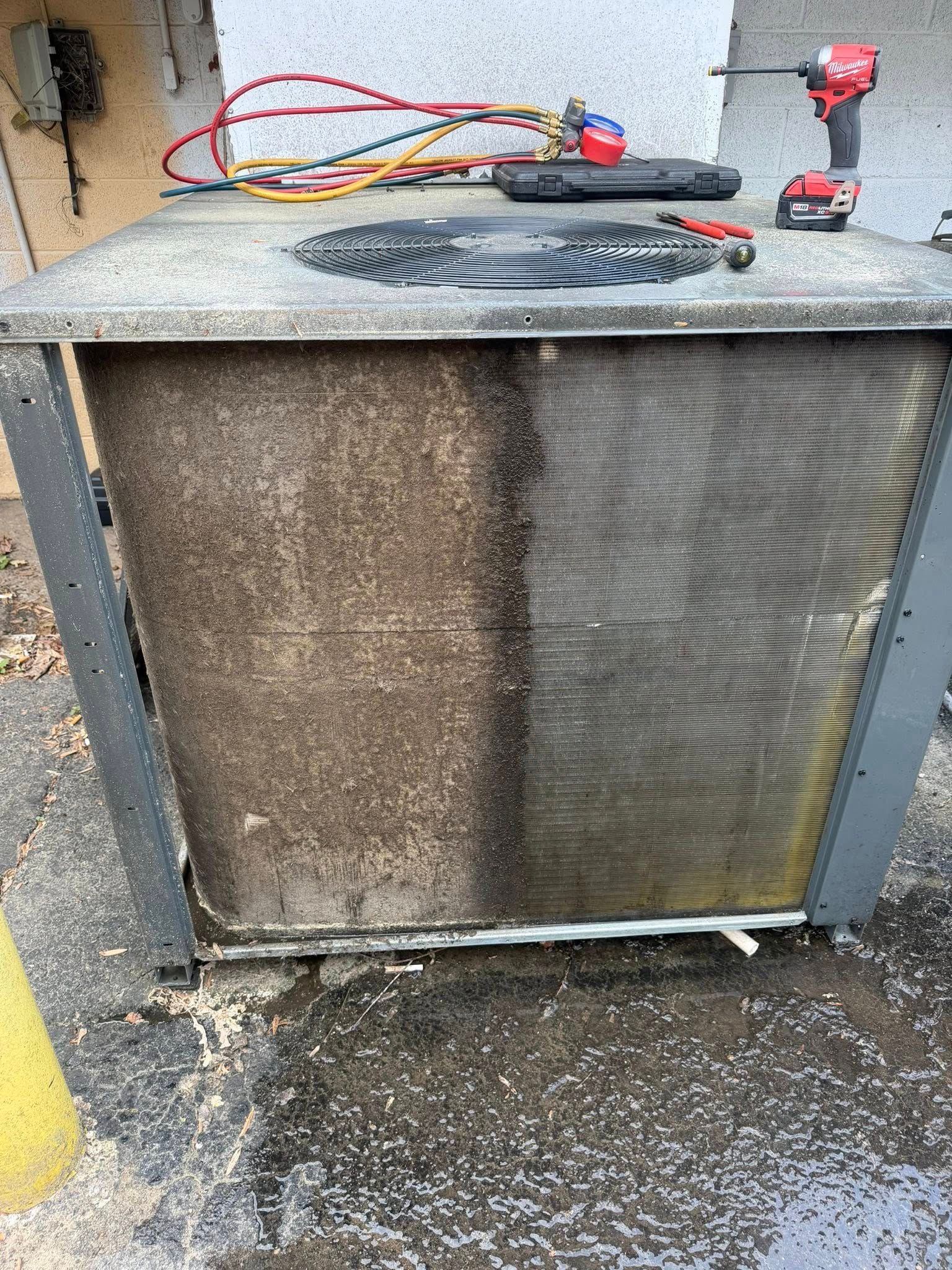 Outdoor HVAC unit with a dusty top holding a tool case, tangled cords, and a red cordless drill