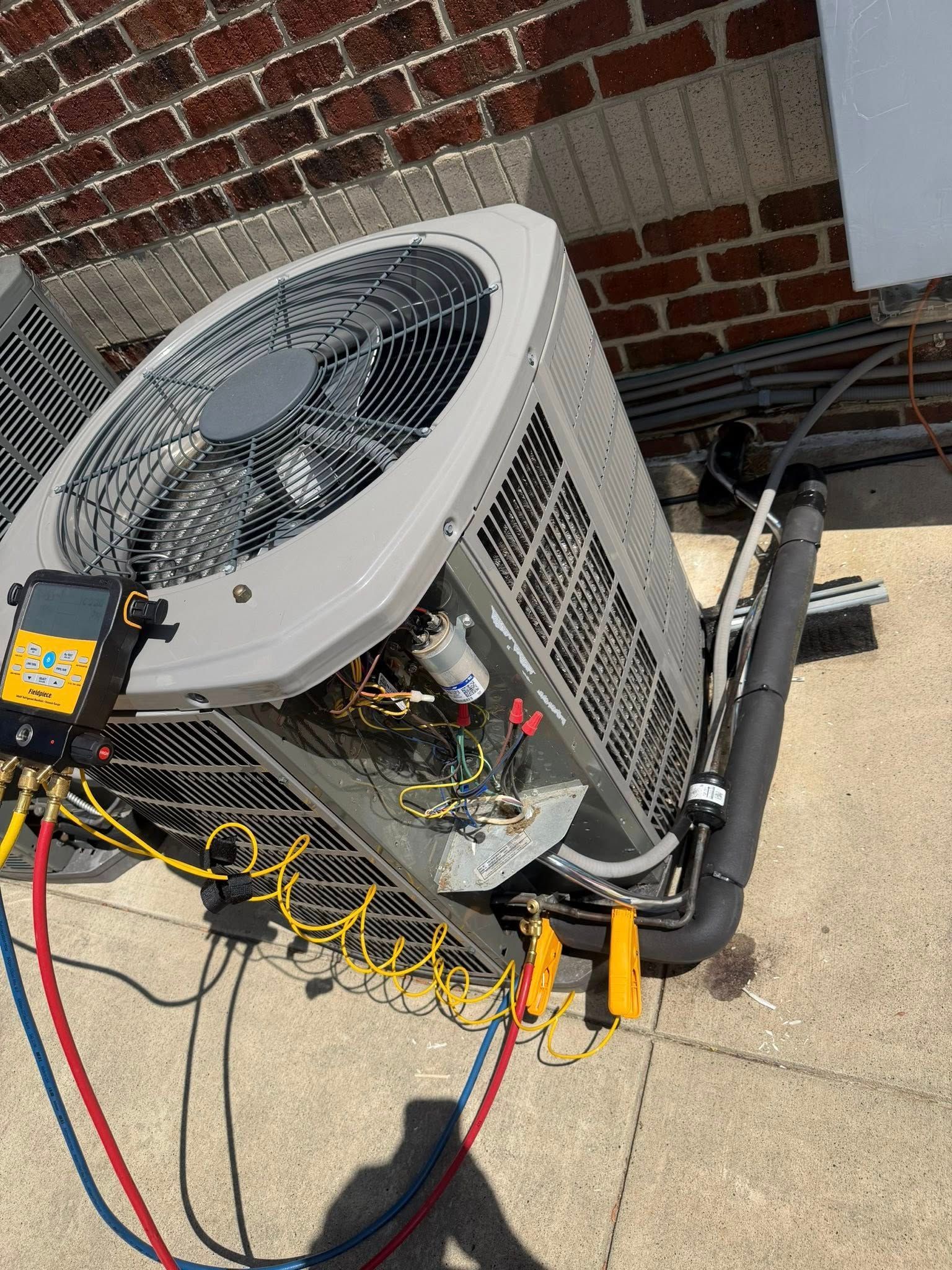 Outdoor HVAC unit with exposed wiring and hoses beside a brick wall and concrete ground