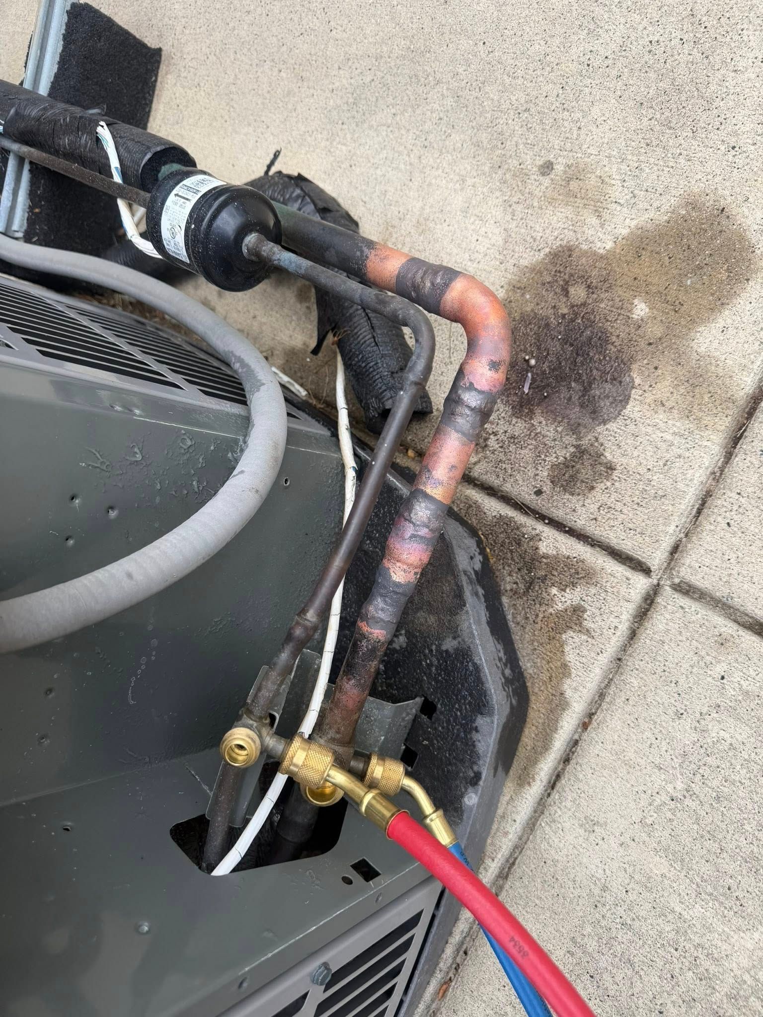 Close-up of HVAC unit refrigerant pipes with red and black hoses attached and rusted copper tubing on concrete.