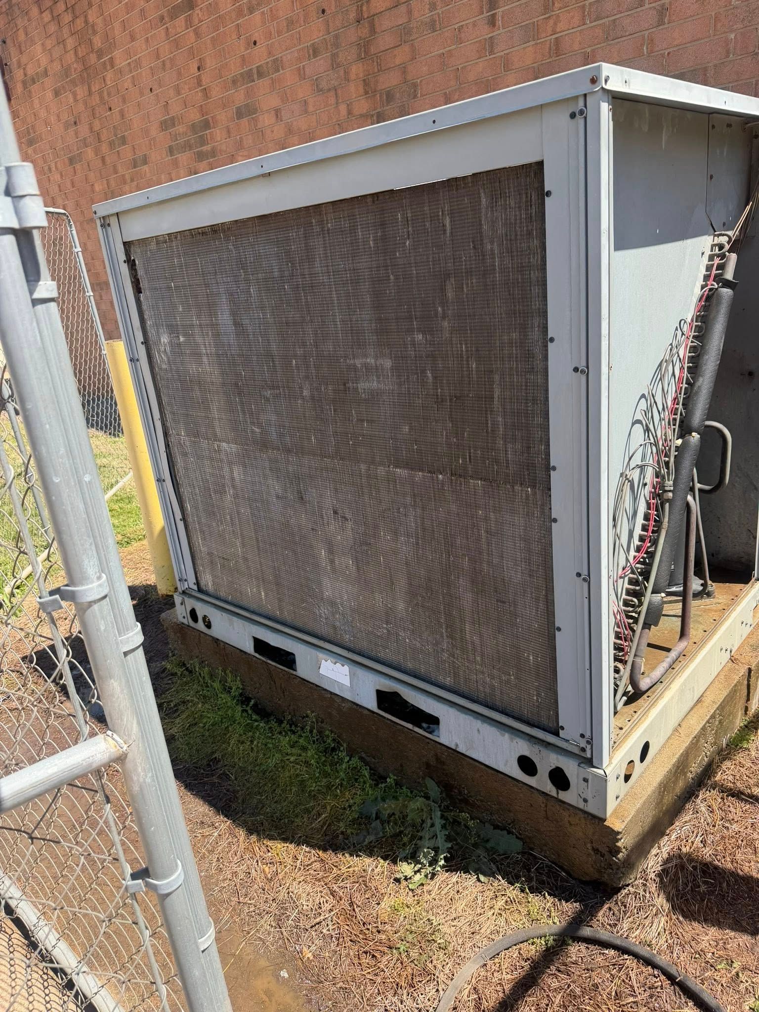 Large outdoor HVAC unit with a rusted coil panel, beside a chain-link fence and brick wall.