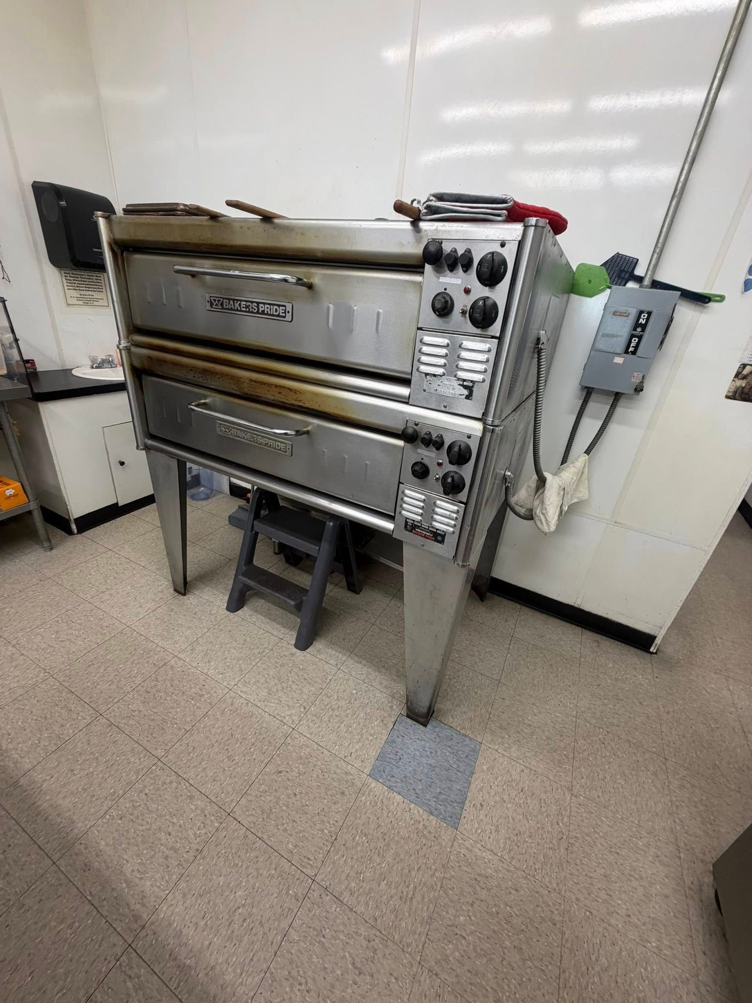 Stainless steel commercial oven in a tiled kitchen, with control knobs and a stool in front.