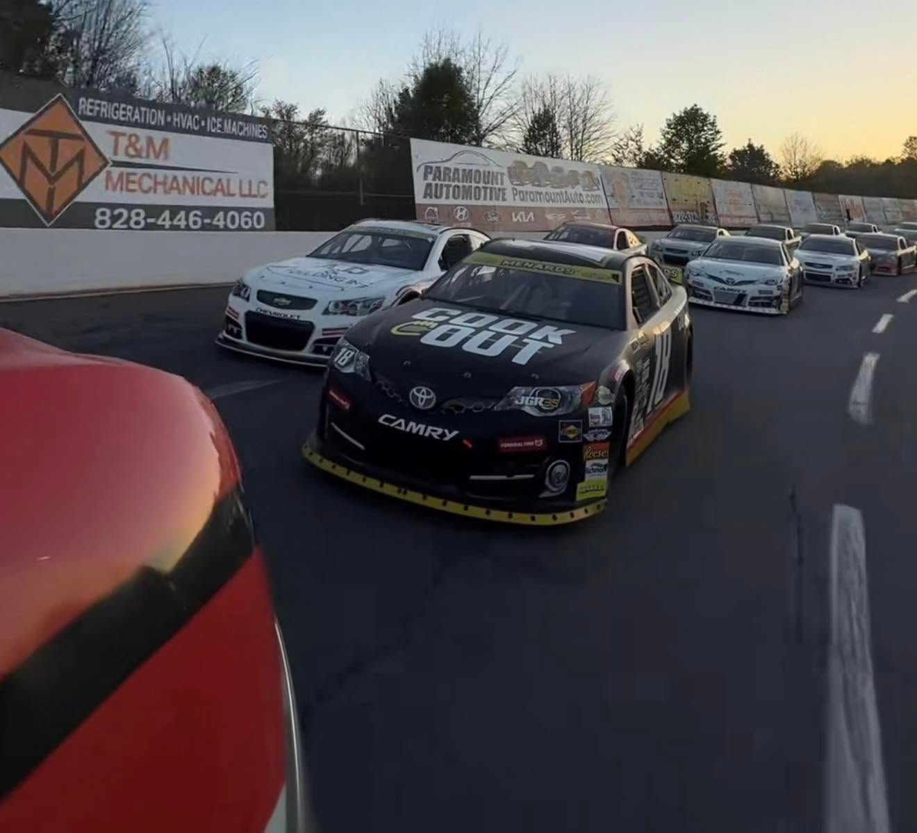 Race cars lined up on a street, front black #18 car beside a red car at sunset.