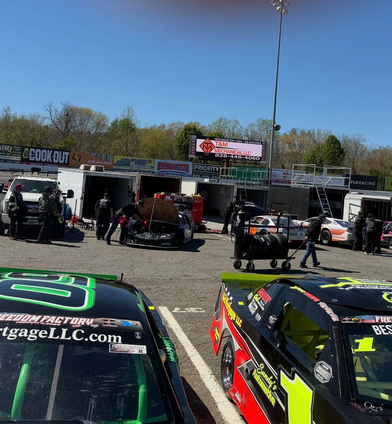Racing pit lane with stock cars and crew under a clear blue sky at a track.