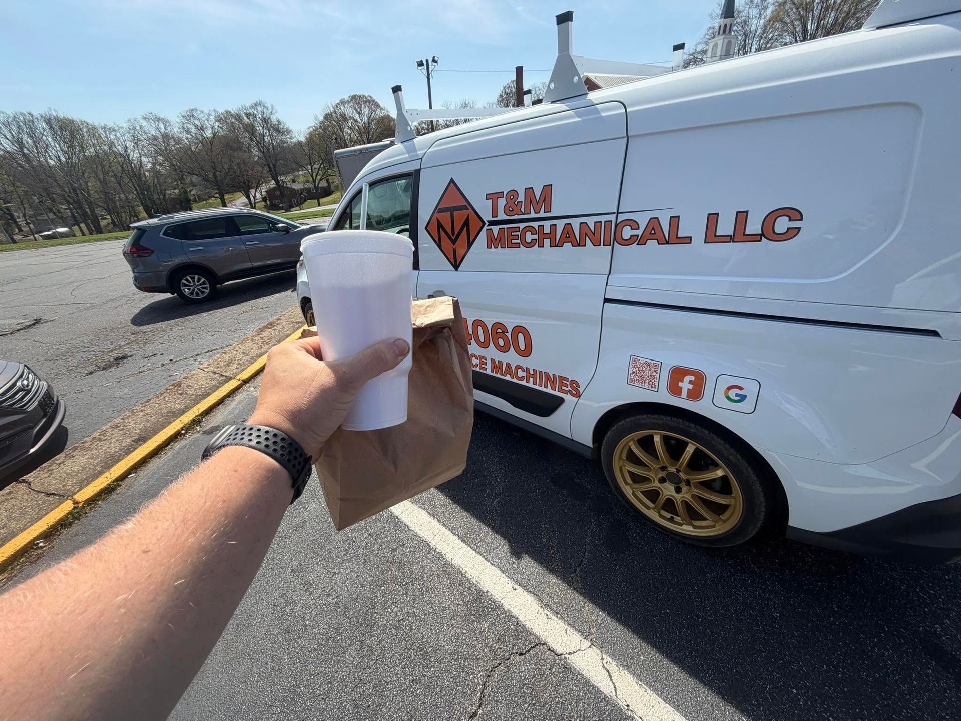Hand holding a drink and bag beside a white Tim Mechanical LLC van in a parking lot.