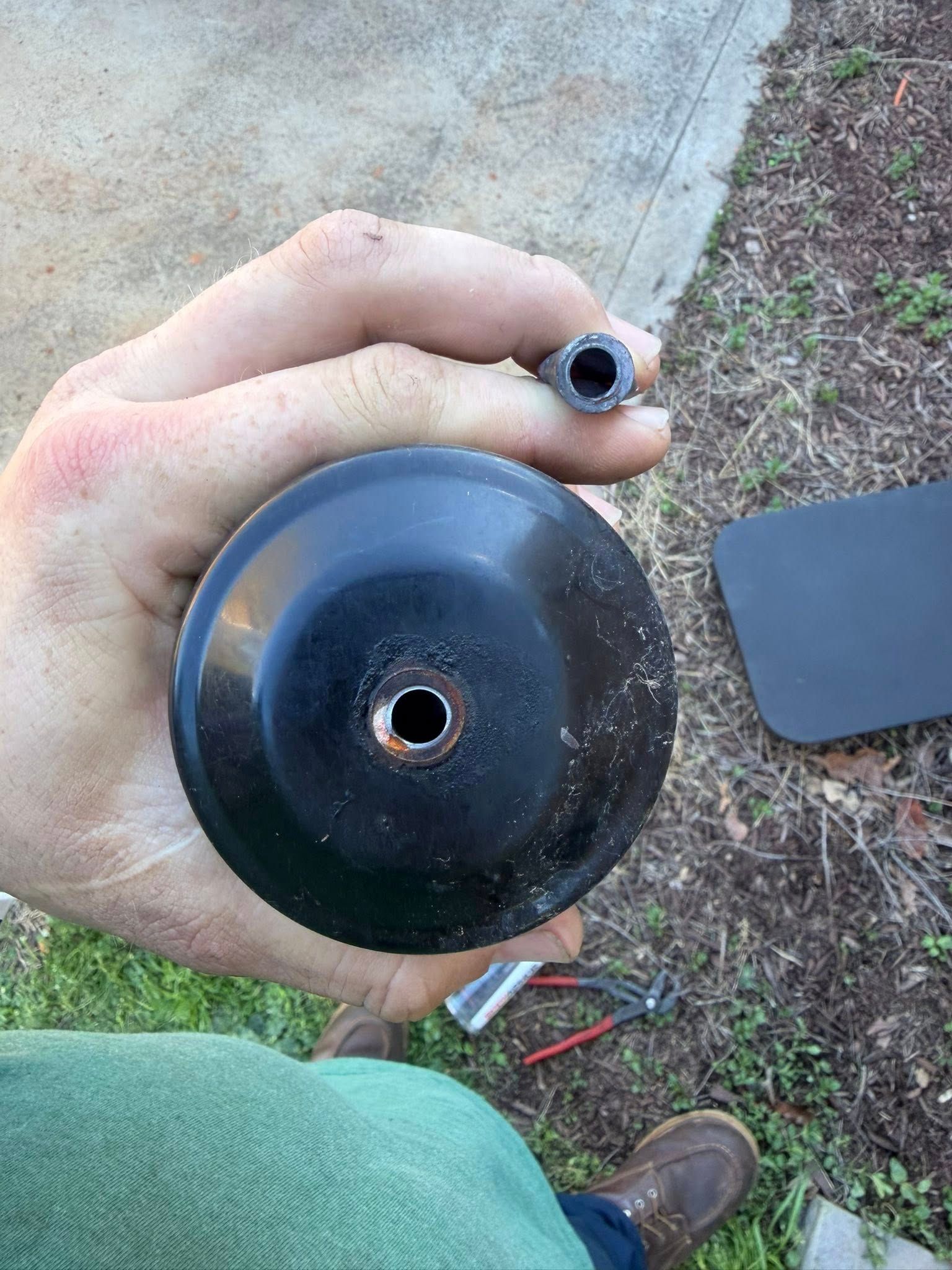 Hand holding a black circular part with a center hole outdoors on pavement and grass.