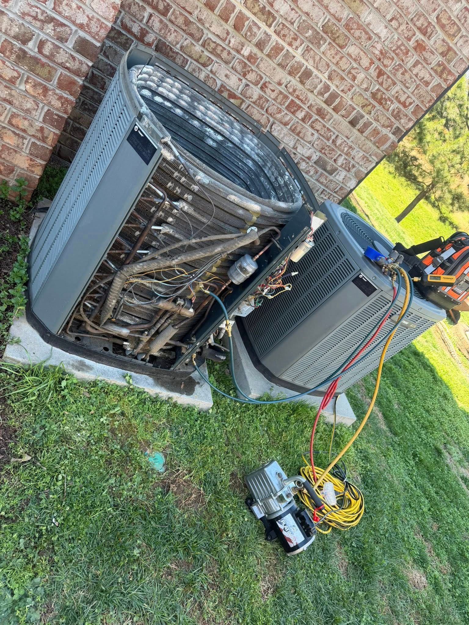 Damaged outdoor HVAC unit beside brick wall, with tools and wires on the grass.