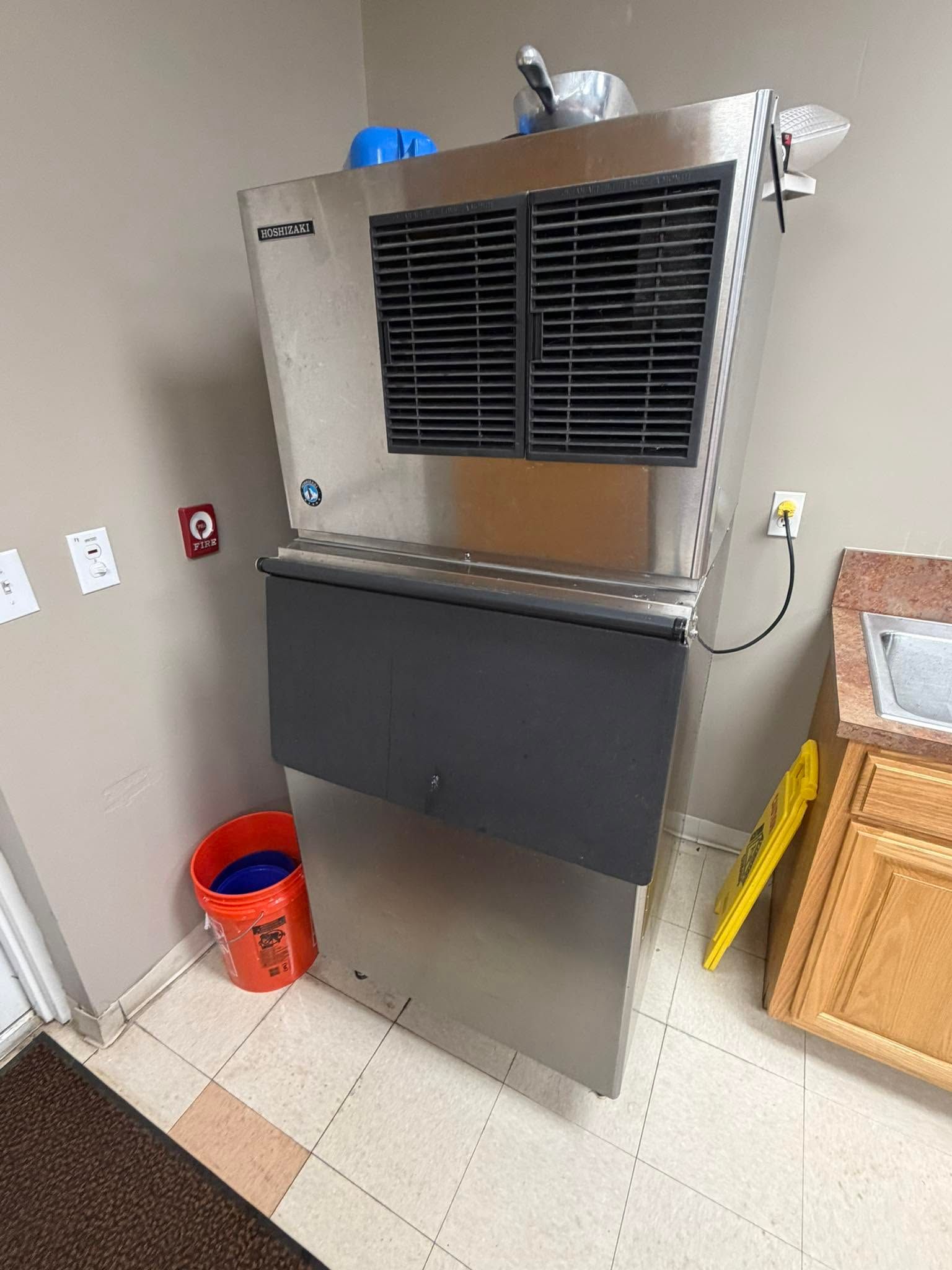 Stainless steel commercial ice machine in a kitchen corner with a black front bin and a red bucket nearby