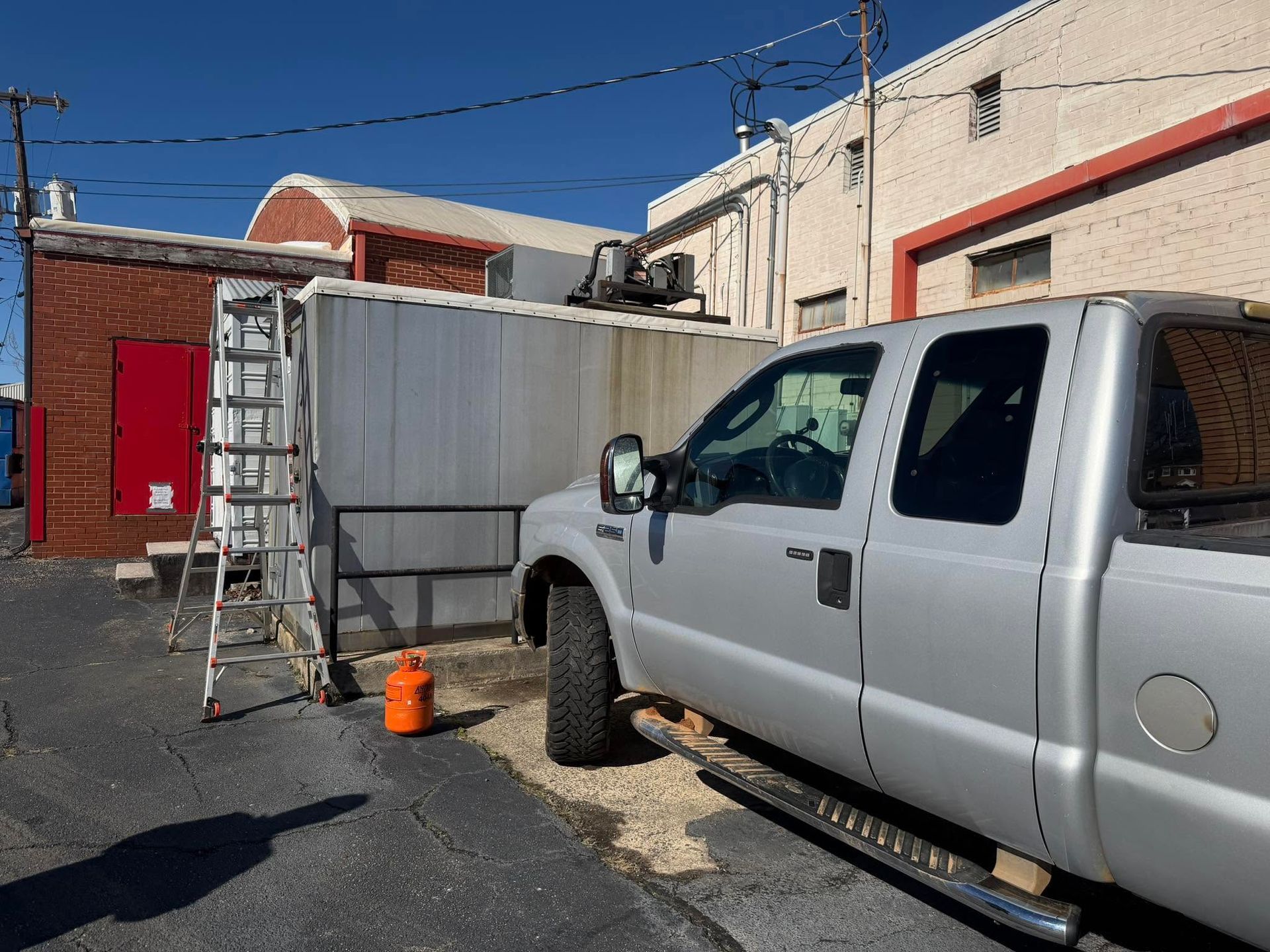 Silver pickup truck beside a white container in an alley, with a ladder and orange cone nearby.