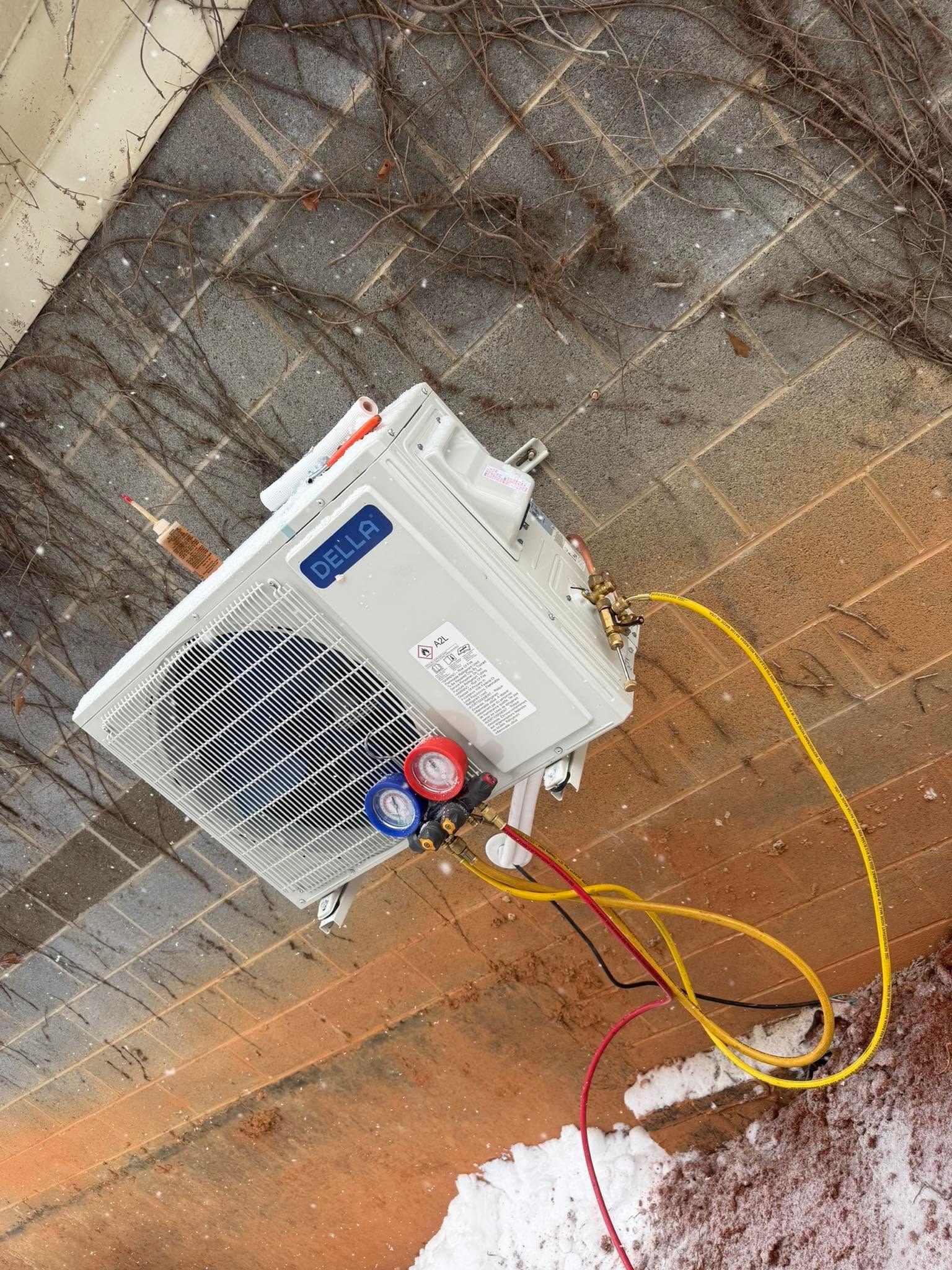 White outdoor AC unit with yellow hoses on a dirt and brick wall, with a red valve gauge attached