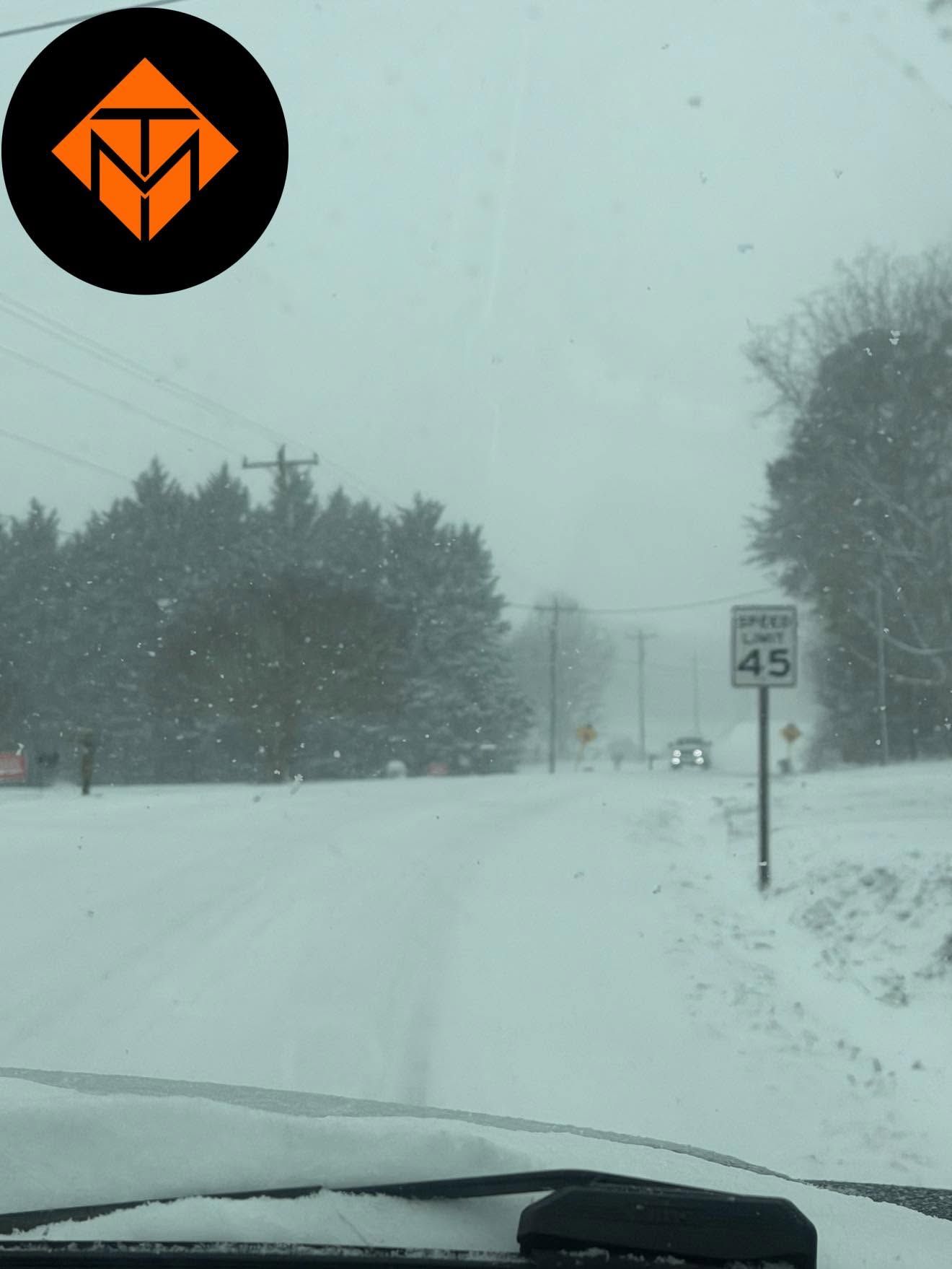Snow-covered road in a snowstorm with a 45 mph speed limit sign and trees lining the roadside