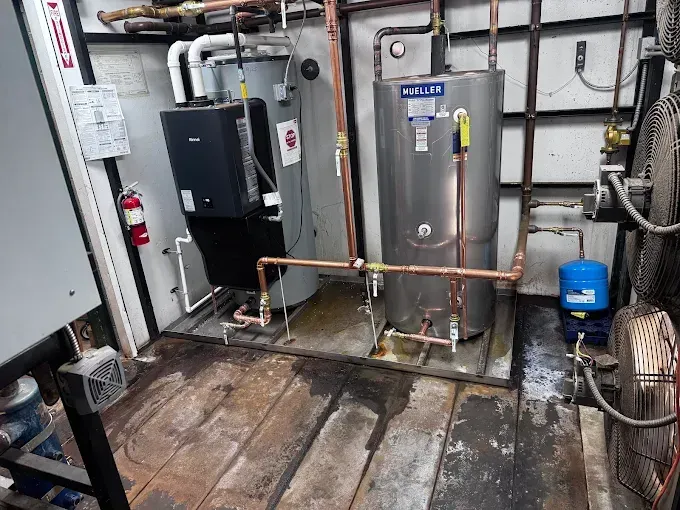 Utility room with boiler, water heater, pipes, and a floor fan on a worn concrete floor.