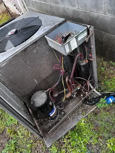 Outdoor air conditioner unit with exposed wiring and open control panel beside a wall.