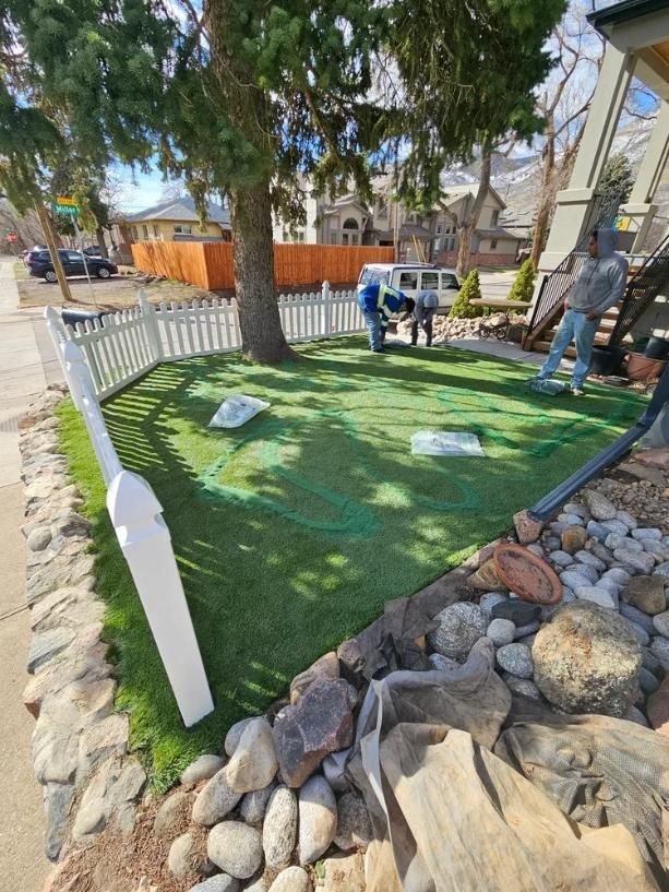 Workers installing artificial turf in a front yard, enclosed by a white picket fence and surrounded by rocks.