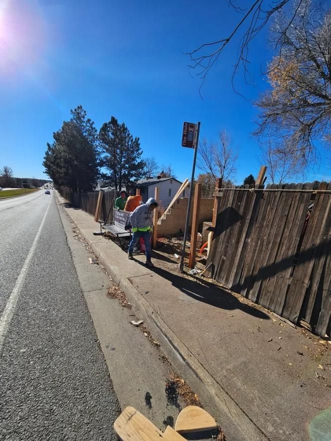 Two people repairing a wooden fence alongside a road. 