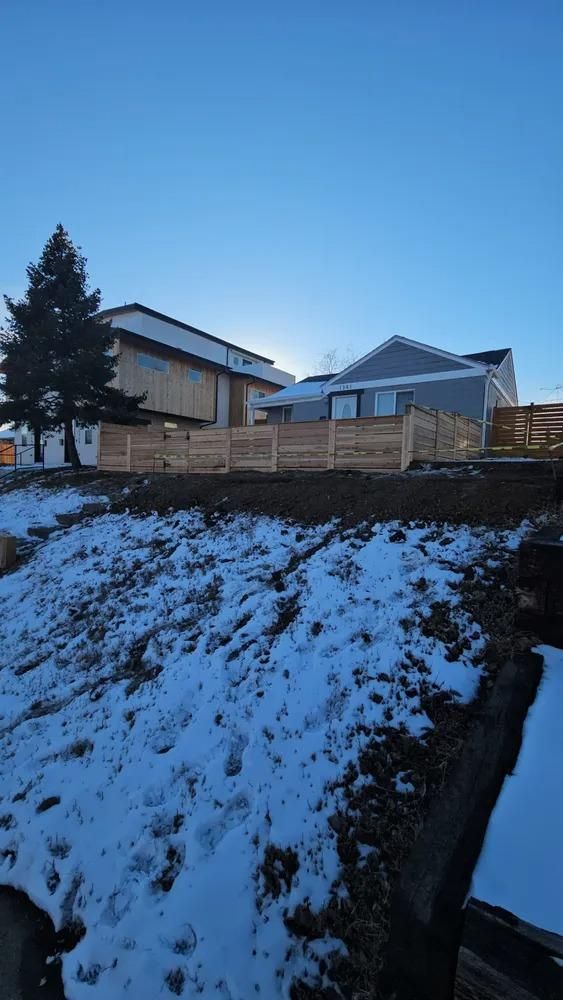 Snowy hillside with wooden fence, houses, and a tree against a clear, blue sky.