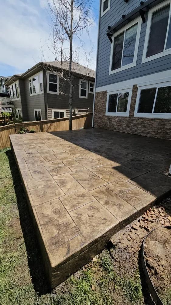 Stamped concrete patio in backyard of a two-story home, with wooden fence and surrounding grass.