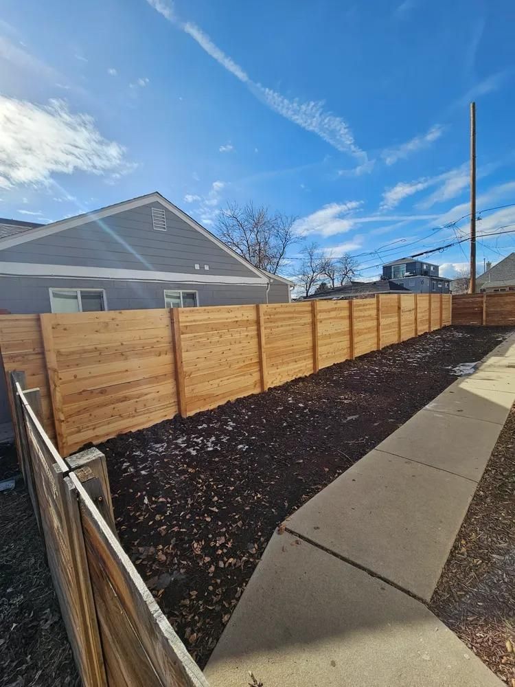 Wooden fence bordering a yard with dark mulch along a sidewalk, under a blue sky.