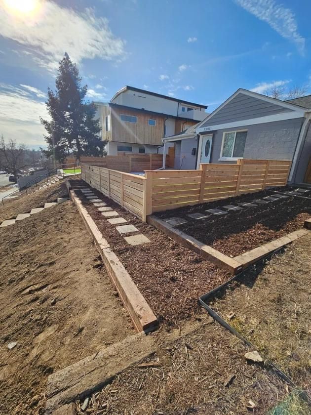 Wooden fence surrounding a yard with mulch, stepping stones, and a house under a blue sky.