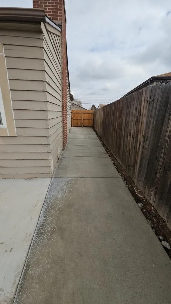 Concrete walkway alongside a tan house and a wooden fence, leading to a gate under a cloudy sky.