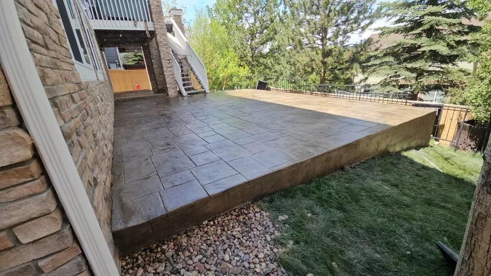 Stamped concrete patio next to a stone wall and deck, overlooking a grassy yard with trees.