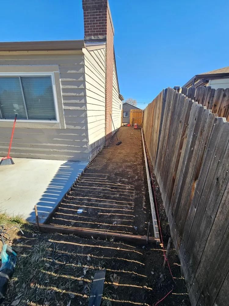Narrow backyard pathway alongside a fence and house, with exposed soil and construction debris.