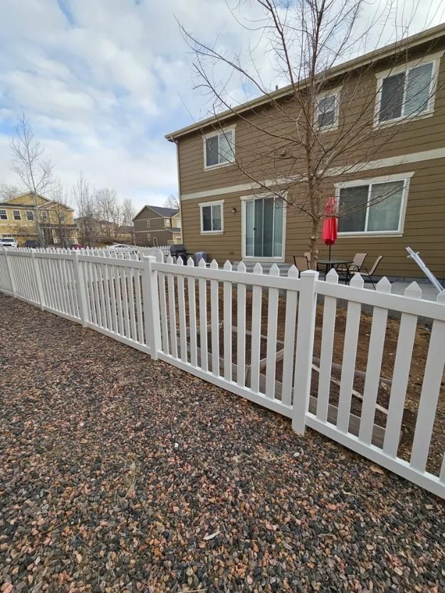 White picket fence surrounds a backyard with a two-story tan house, patio, and gravel ground.