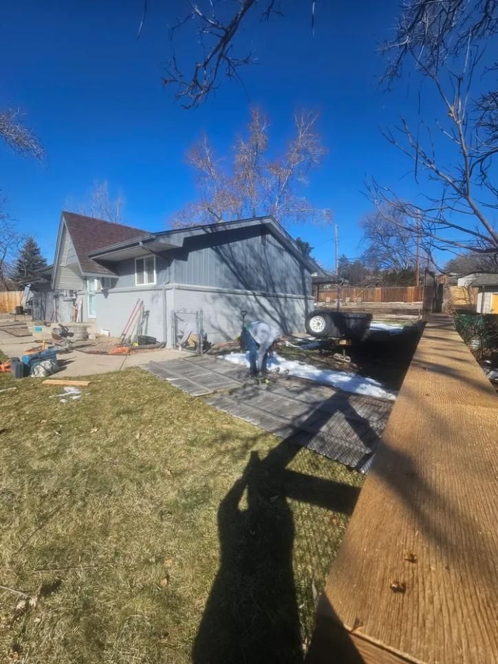 A person working on paving stones next to a house with blue siding, against a clear blue sky.