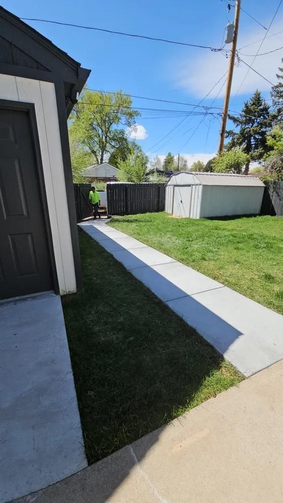Backyard with concrete path, garage on left, shed and fence in background, person mowing.