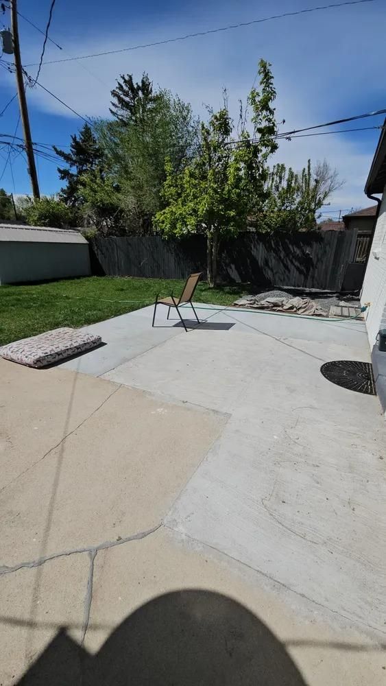 Backyard patio with concrete and grass, chair, shed, trees, and blue sky.