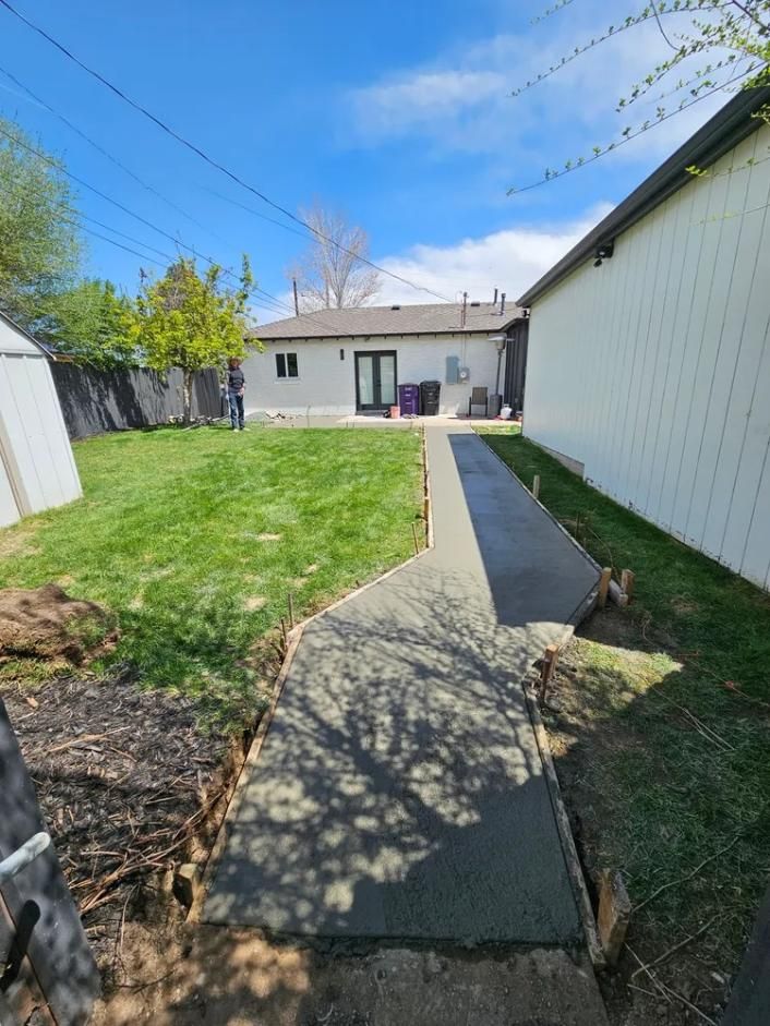 Newly poured concrete walkway in a backyard, beside a house and green lawn, blue sky overhead.