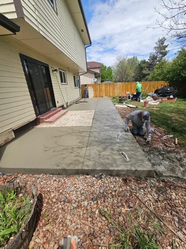 Workers pouring and finishing concrete patio next to a house with a sliding glass door and fenced backyard.