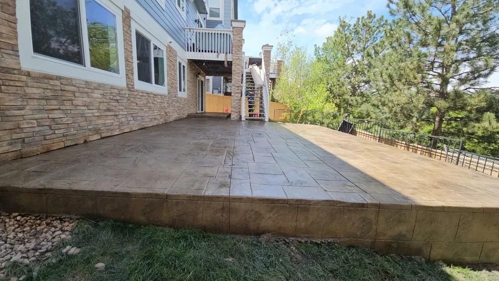 Concrete patio next to a house with a stone facade, grassy lawn, and trees under a cloudy sky.