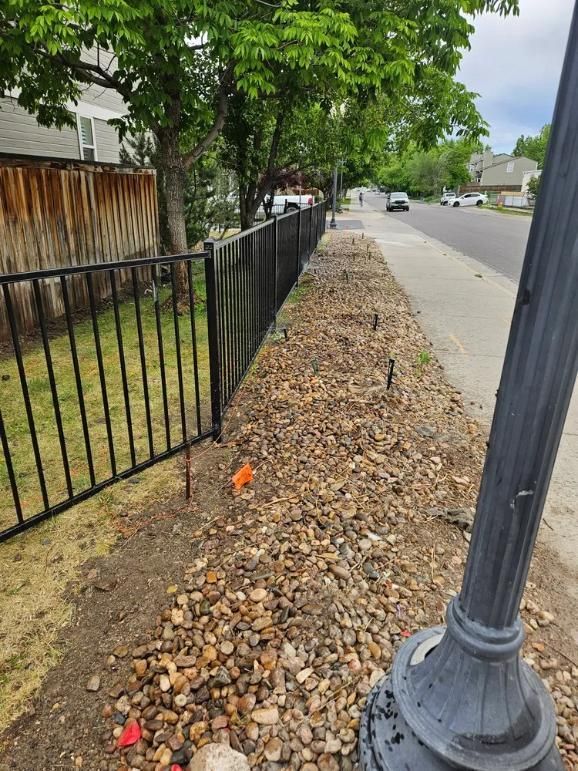 Black metal fence alongside a sidewalk with gravel and a street in the background. 