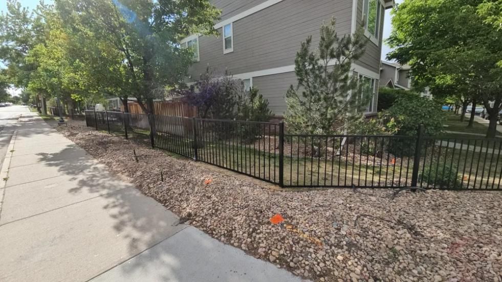 Black metal fence along a sidewalk in front of a house, landscaping with mulch and trees.